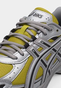 Κοντινό πλάνο κίτρινου και ασημί παπουτσιού τρεξίματος Asics με γκρι κορδόνια και λογότυπο της μάρκας με μαύρες λεπτομέρειες στη πλευρά.