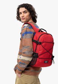 Un sac à dos rouge avec un design structuré, comportant une poche en mesh et des accents noirs. Porté sur l'épaule, associé à un pull à motifs.