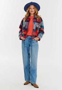 Pestrý károvaný kabát v modrých, červených a zelených odtieňoch, spárovaný s oranžovým svetrom, širokými džínami a hnedými lodičkami.