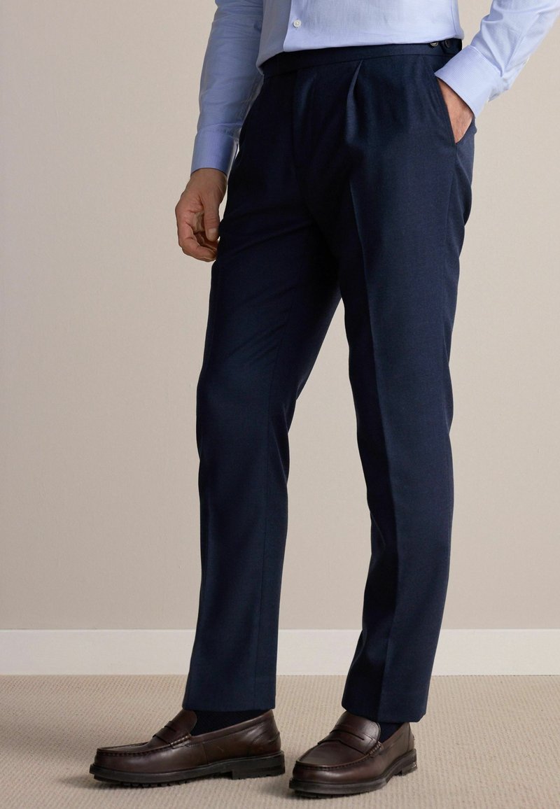 Pantalons taillés bleu marine avec une texture lisse, présentant des plis et des poches latérales. Associés à des mocassins en cuir marron.