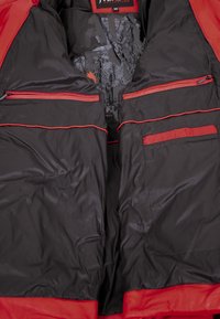 Schwarze Jackeninnenseite mit leuchtend roten Akzenten, ausgestattet mit mehreren Reißverschlusstaschen und einer eleganten, glatten Textur. Muster auf dem Futter sichtbar.