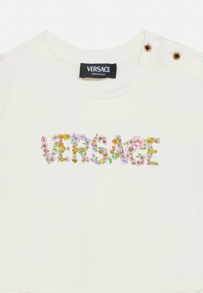 Λευκό πουκάμισο Versace με κείμενο "VERSACE" σε σχέδιο με λουλούδια στο στήθος και δύο κουμπιά σε χρυσό χρώμα στον αριστερό ώμο.