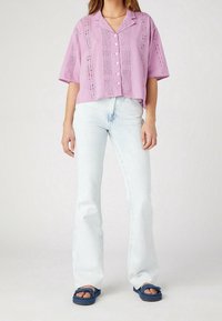 Osoba ubrana w lawendową bluzkę z krótkim rękawem zapinaną na guziki, z ażurowego materiału, jasnoniebieskie dżinsy z wysokim stanem i granatowe sandały na odkryte palce.