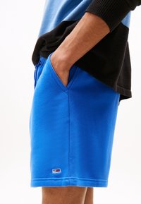 Modré bavlněné kraťasy s páskem na šňůrku, bočními kapsami a detailem loga na lemu. Měkká textura, pohodlný design.