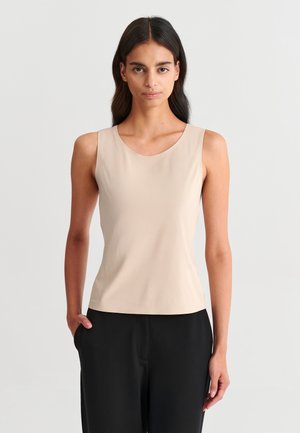 Femme aux longs cheveux foncés portant un haut beige sans manches et un pantalon noir, debout devant un fond clair uni.