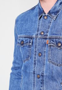 Klasická modrá džínová bunda s vybledlým efektem, měděnými knoflíky, dvojitými kapsami na prsou a oranžovou značkou Levi's na levé kapse.