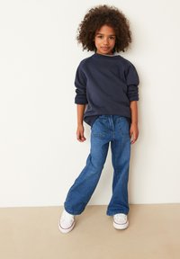 Marineblauwe sweatshirt met opgerolde mouwen, gecombineerd met blauwe flared jeans en witte sneakers met rode accenten. Glad materiaal, casual ontwerp.