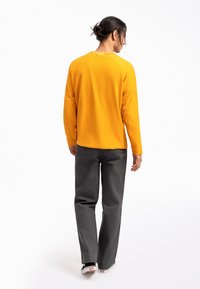 Camicia a maniche lunghe gialla con scollatura rotonda e spacchi laterali, abbinata a pantaloni grigio scuro. Il materiale sembra morbido con una texture semplice.