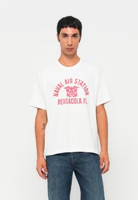 Jaunas vyras dėvi baltą marškinėlį su raudonu užrašu "Jūrų oro stotis Pensacola, FL" ir emblema, derinantį su mėlynais džinsais, stovi prieš baltą foną.