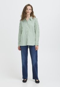 Giovane donna che indossa una camicia a righe verticali verdi e bianche, jeans blu e scarpe nere, in piedi contro uno sfondo bianco semplice.