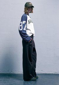 Cămiză cu mânecă lungă, albă, cu mâneci bleumarin și text verde. Pantaloni întunecați, lejeri. Căciulă verde cu logo. Texturi netede.