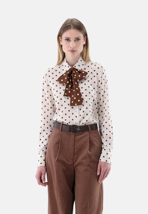 Donna che indossa una camicetta bianca a pois marroni e un grande fiocco marrone, abbinata a pantaloni marroni a vita alta e una cintura scura.