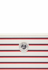Portefeuille zippé blanc avec des rayures verticales rouges et navy. Présente un logo rond avec le texte "Roland Garros" et des accents noirs contrastants. Surface texturée.
