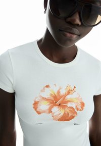 Hvid bomulds t-shirt med orange hibiscus tryk på forsiden, rund halsudskæring og korte ærmer. Glat tekstur, tætsiddende design.