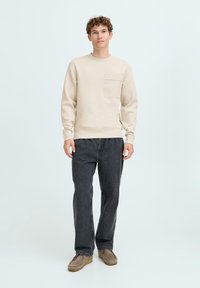 Beige sweatshirt met een voorzak, gecombineerd met donkergrijze jeans en bruine suède instap schoenen, staand tegen een lichte achtergrond.