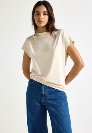 Giovane donna in piedi con le mani dietro la schiena, indossa una maglietta beige larga e jeans blu a vita alta su uno sfondo bianco semplice.