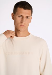 Beige sweatshirt med ribbad rundhals, med präglad text "COPENHAGEN LINDENBERGH" på framsidan. Mjuk textur, avslappnad design.