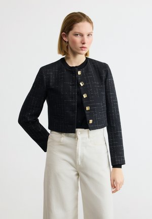 Femme portant une veste courte noire à boutons dorés et un pantalon blanc taille haute, debout devant un fond blanc uni.