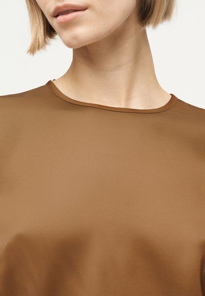 Γυναίκα με κοντά μαλλιά που φοράει ένα καφέ τοπ με στρογγυλή λαιμόκοψη, εμφανίζοντας τον λαιμό και μέρος του προσώπου της μπροστά σε ανοιχτό φόντο.