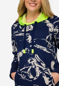 Sweatshirt bleu marine avec un imprime graphique beige, doté d'une doublure de capuche vert fluo, de cordons de serrage ajustables et d'une poche frontale.