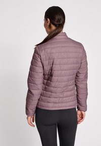 Lys lilla dunjakke med quiltet tekstur, høj krave og lange ærmer, parret med sorte leggings. Bagfra ses den tætsiddende design.