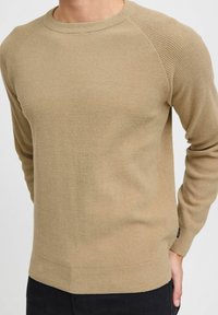 Beige, getextureerde gebreide trui met ronde halslijn en raglanmouwen. De zoom is recht, en de stof heeft een subtiel, organisch patroon.