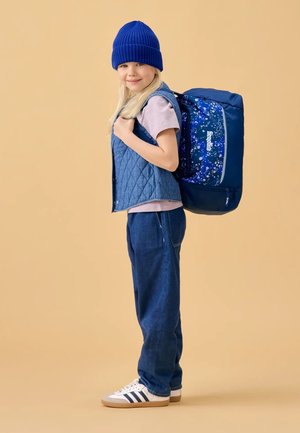 Blue quilted vest over a light pink t-shirt, blue denim pants, and white sneakers with navy stripes. A blue patterned backpack rests on the shoulder.