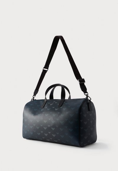 Σκούρο μπλε σακ βουαγιάζ με επαναλαμβανόμενο μοτίβο λογότυπου αετού, διπλές επάνω λαβές και ρυθμιζόμενο μαύρο ιμάντα ώμου, τοποθετημένο σε απλή φόντο.