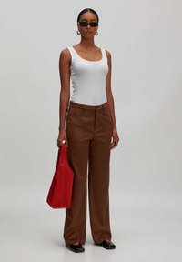 Braune weit geschnittene Hosen mit glatter Textur, kombiniert mit einem eng anliegenden weißen Tanktop. Die Person hält eine rote Tote-Bag und trägt schwarze Sonnenbrillen.