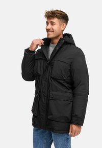 Veste noire rembourrée avec capuche, deux grandes poches avant et fermeture éclair. Le matériau semble lisse et résistant à l'eau.