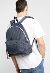 Mörkblå rutig tygryggsäck med en framficka och vit dragkedja. Har justerbara axelremmar och en tagg med varumärket.