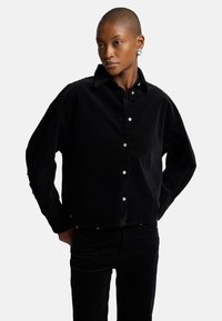 Camicia di corduroy nera con colletto, bottoni frontali e maniche lunghe. Il tessuto presenta una consistenza morbida con una forma leggermente oversize.
