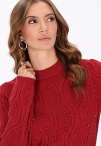 Czerwony sweter z dzianiny z teksturowanym wzorem z wypukłymi owalnymi kształtami i drobnymi ozdobami. Wysoki dekolt i prążkowane mankiety.