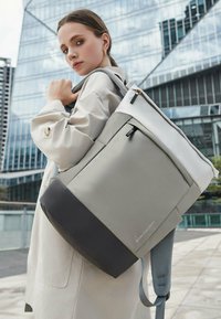 Un sac à dos gris et noir avec une finition lisse, comportant une fermeture éclair supérieure, une poche latérale et des bretelles texturées. Porté sur l'épaule.