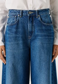 Wide-leg blue denim jeans with a faded finish, featuring a button closure, belt loops, and front pockets, displayed against a light background.