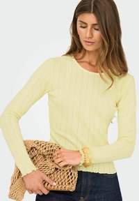 Ljusgul ribbad långärmad topp med rundad halsringning. Texturerat tyg och vågig nederkant. Accessoar: en beige nätad clutchväska.