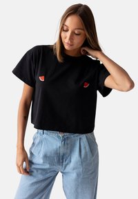 Czarna, krótkowłosa koszulka z bawełny, z małymi, haftowanymi wzorami arbuza z przodu. W parze z jasnoniebieskimi, wysokimi dżinsami.