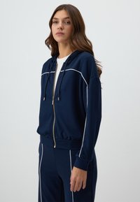 Navy zip-up hoodie with white piping. Features a drawstring hood, ribbed cuffs, and side pockets. Soft texture, fitted design.