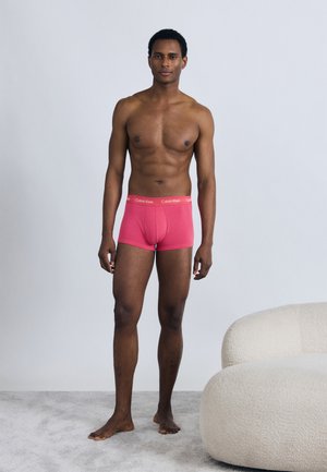 Sarkani kokvilnas bokseršorti ar elastīgu Calvin Klein jostu, valkāti uz kailas vīrieša figūras neitrālā vidē.