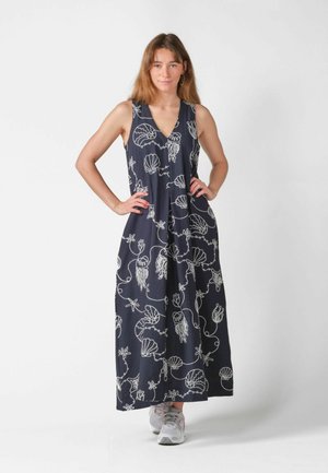 Frau steht mit den Händen in der Hüfte, trägt ein ärmelloses marineblaues Maxikleid mit weißen Mustern von Meerestieren und Muscheln sowie graue Sneakers.