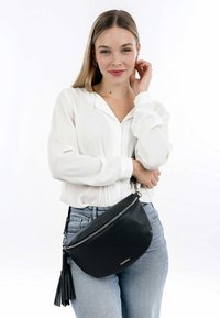 Zwarte leren schoudertas met een gestructureerd oppervlak, ritssluiting en een decoratieve franjes detail. Casual gedragen over een wit shirt.
