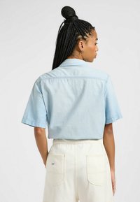Camisa de manga corta azul claro con cuello, que presenta costuras en el yugo trasero y un corte relajado, combinada con pantalones cortos crema.