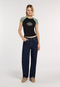 Donna in piedi, rilassata, indossa una camicia corta nera e verde con il logo "Dickies", jeans blu scuro e scarpe beige con lacci, su uno sfondo bianco.