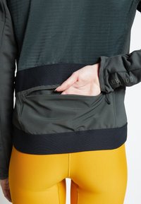 Main atteignant la poche arrière zippée d'une veste vert foncé portée avec des leggings jaune vif.