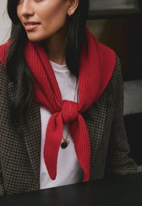 Sciarpa rossa a maglia annodata davanti, sovrapposta a una camicia bianca, abbinata a un blazer a quadri. Presenta una collana con pendente rotondo.