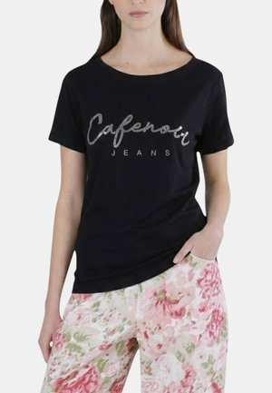 Donna che indossa una t-shirt nera "Cafenoir Jeans" e pantaloni rosa a fiori, con una mano in tasca su sfondo semplice.