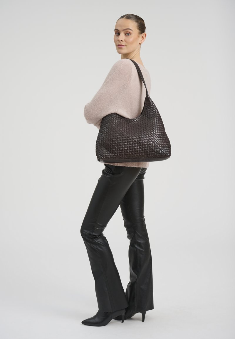 Žena v béžovém svetru a černých kožených kalhotách stojí bokem a na rameni nese velkou černou tkanou kabelku.