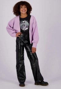 Nasmeešena djevojka stoji s rukom na bokovima, nosi ljubičasti kardigan, crnu majicu s grafičkim printom, crne hlače od umjetne kože i crne čizme na sivoj pozadini.