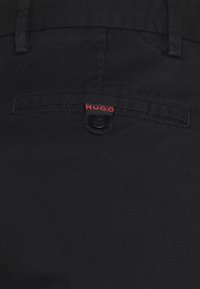 Tessuto nero con una superficie liscia; presenta una tasca sul retro con il logo rosso "HUGO" e una chiusura a bottone. Design semplice e minimalista.