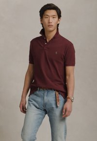 Νεαρός άνδρας φορεί μπλουζάκι polo μπορντό, ανοιχτού μπλε τζιν με πλεκτή ζώνη και ρολόι χειρός, στέκεται μπροστά σε απλό ανοιχτό φόντο.